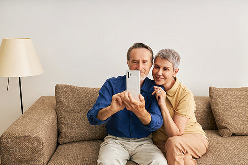 Senior couple in casuals at home sitting on couch taking a selfie