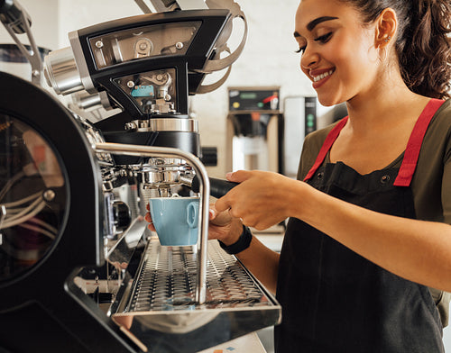 Smiling female barista using coffee maker in cafeteria. Woman in apron preparing espresso.