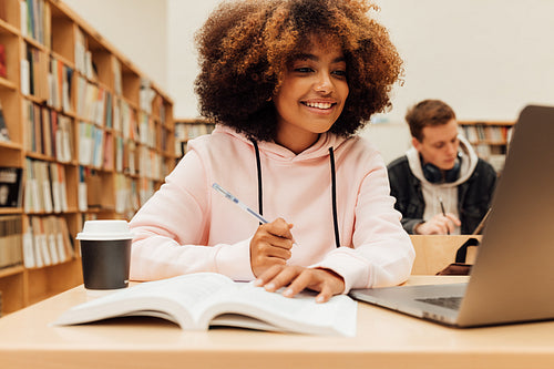 Smiling girl sitting at a table in the library and looking at a laptop