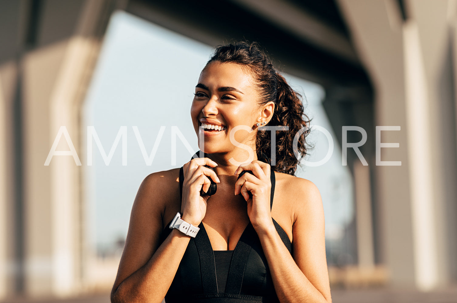 Beautiful happy woman holding headphones and looking away while standing on city street after fitness training	