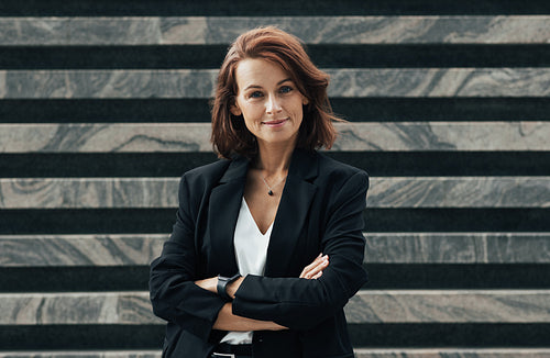 Smiling and confident middle-aged female with crossed hands. Businesswoman in black formal clothes with ginger hair.