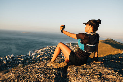 Young woman in sportswear relaxing after hike and making selfie while sitting on rock