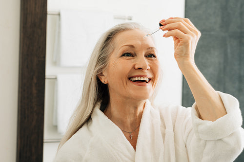 Smiling senior woman holding pipette with hyaluronic acid. Happy female applying liquid facial treatment.