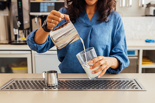 Mid section of a female barista pouring milk from steel frothing pitcher into a glass