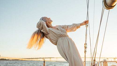 Side view of a mature woman with long grey hair holding a rope on a private yacht and enjoying sunset