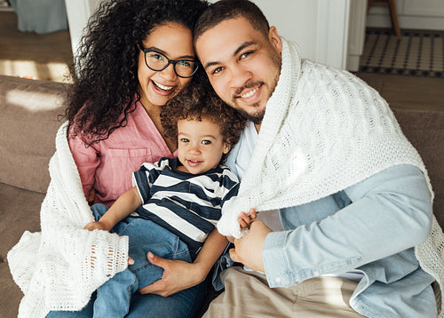 Mother, father, and their cute son looking at camera. Family of three sitting at home wrapped in white plaid.