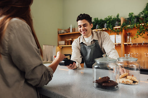 Smiling male barista holds a card machine while the customer pays by NFC. Barista in an apron receiving payment in a cafe at the counter.