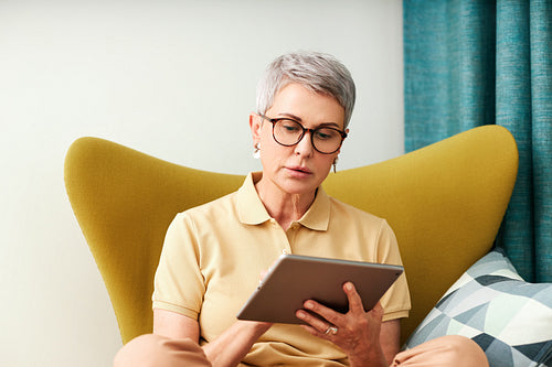 Mature woman sitting at home on armchair using digital tablet. Female in casuals watching content on a tablet.