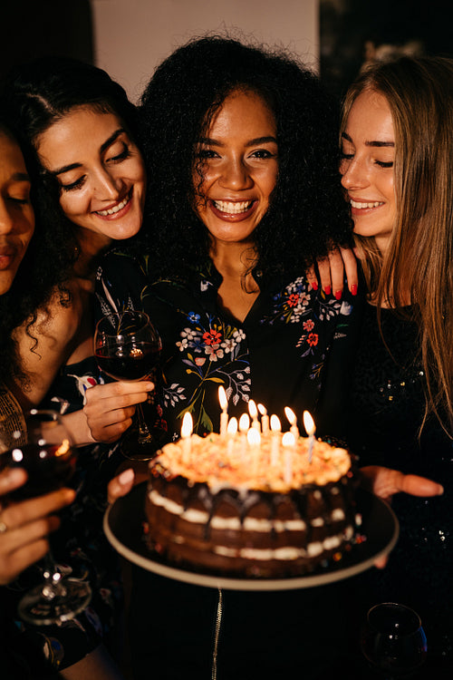 Young happy woman holding a birthday cake, standing indoors surrounded by girlfriends