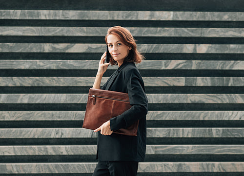 Side view of a confident middle-aged female with a leather folder. Businesswoman with ginger hair making a phone call while standing outdoors.