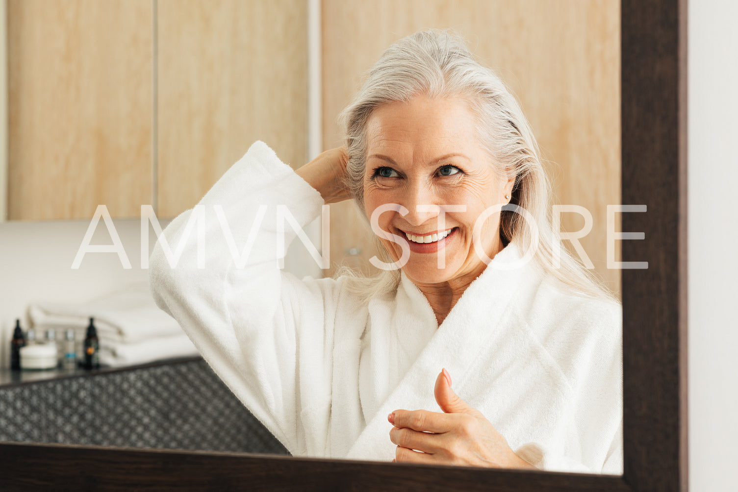 Cheerful aged woman in bathrobe adjusting her grey hair in front of a mirror